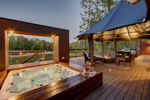 Una terraza de madera con jacuzzi, mobiliario exterior y un área cubierta con una parrilla.