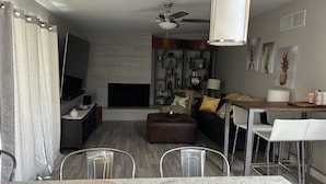Een moderne woonkamer met een open haard, een eethoek met een tafel en stoelen, en een plafondventilator.