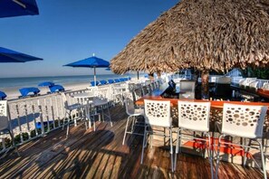 Eine Strandbar mit reetgedeckten Sonnenschirmen, weißen Stühlen und einer Holzterrasse mit Blick auf den Ozean.
