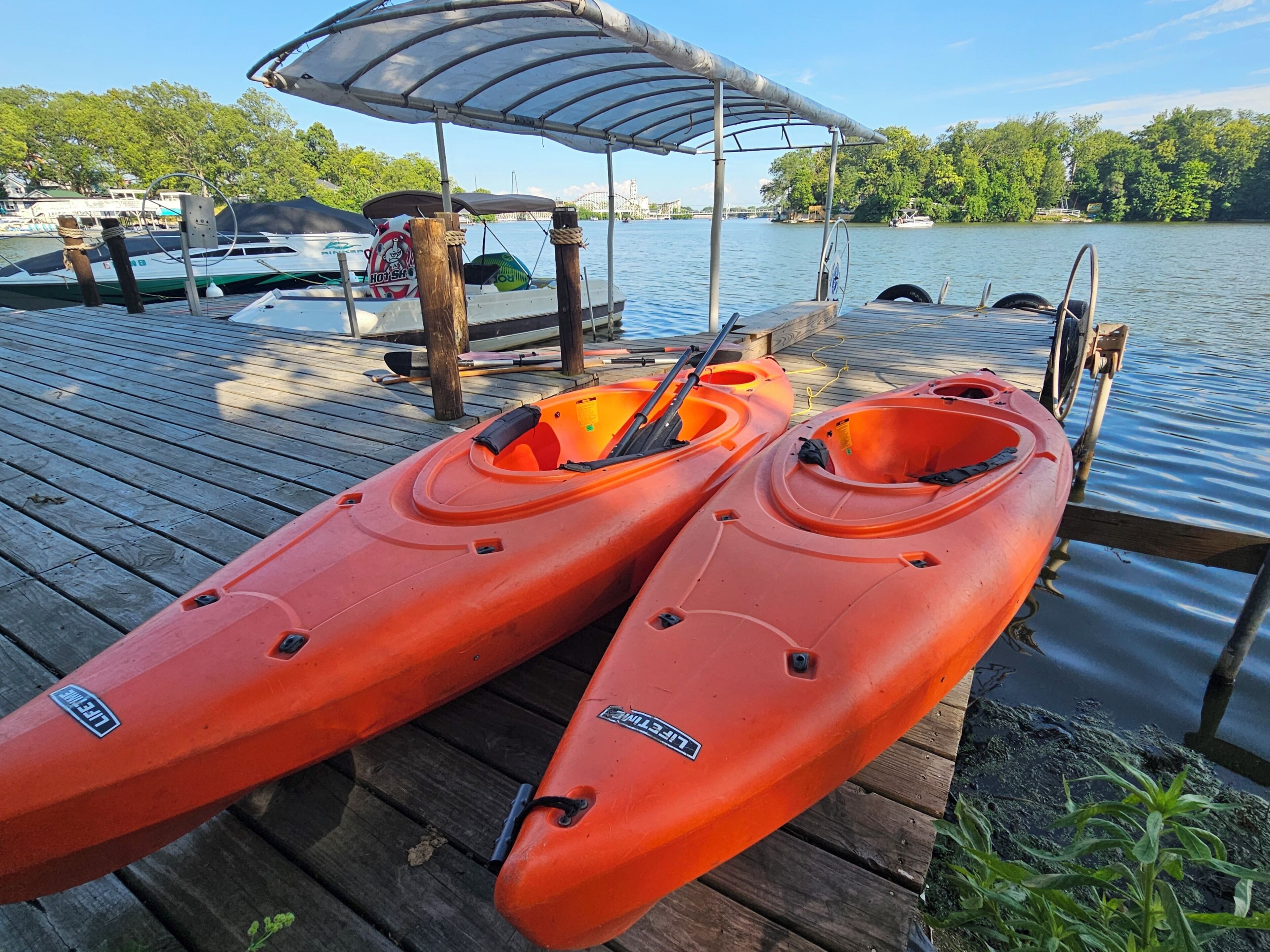 Dos kayaks sobre un muelle de madera con una zona de estar techada, un bote al fondo y un lago rodeado de árboles.