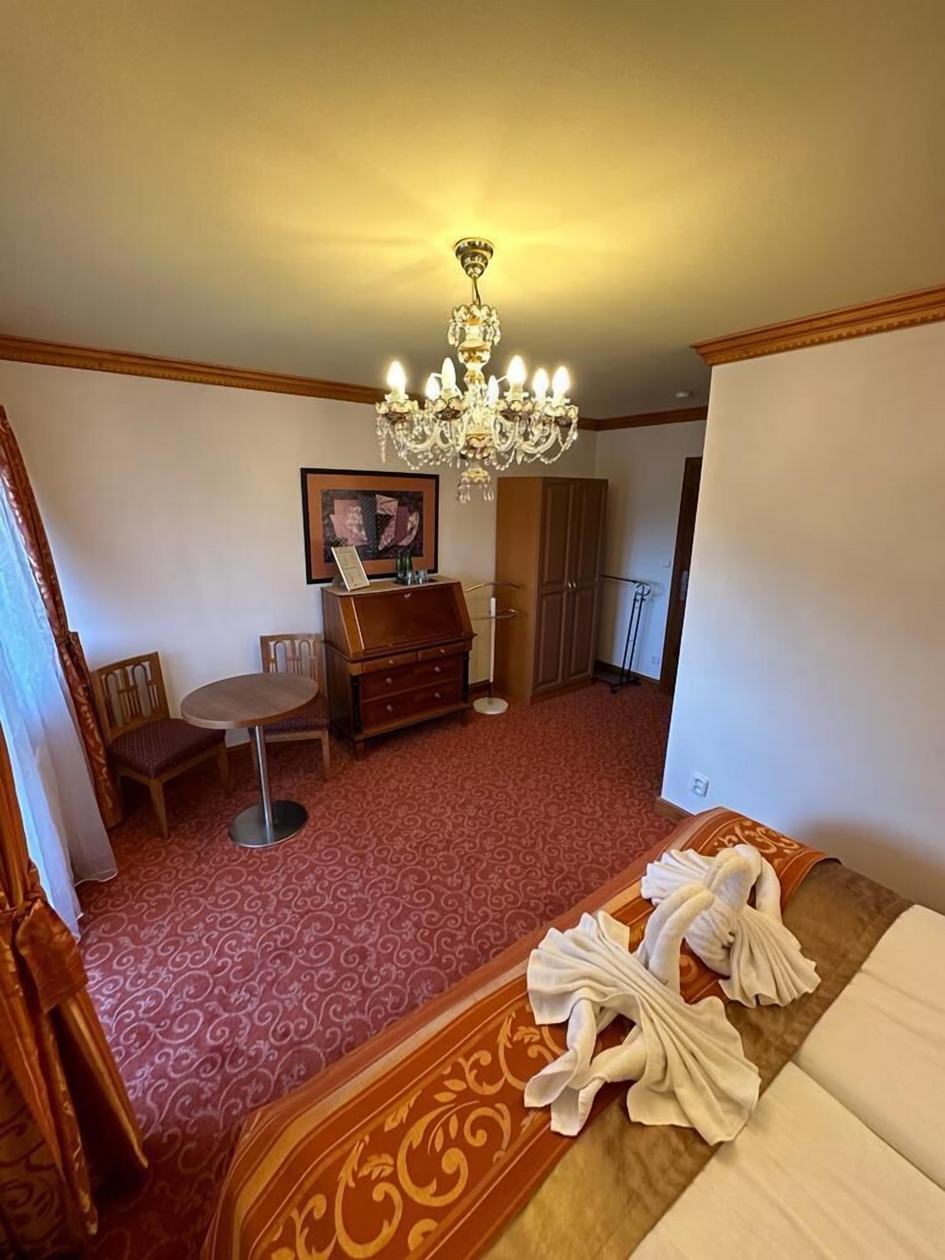 Ein Hotelzimmer mit einem Bett, einem Schreibtisch, einem Stuhl, einer Kommode, einer Chandelüster und einem Gemälde an der Wand.