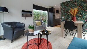 Ein modernes Wohnzimmer mit einem grauen Sofa, einem kleinen runden Tisch, einem roten Teppich, einem Fernseher und einer Küchenzeile mit dunkler Spritzschutzleiste und floralem Wandpapier.