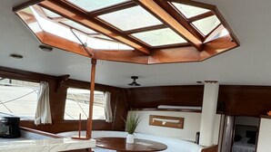 Interior de barco com área de refeições em madeira, sala de estar com almofadas, uma pequena mesa com chapéu e teto com claraboias.