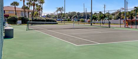 A tennis court with a net, surrounded by palm trees and a fence.