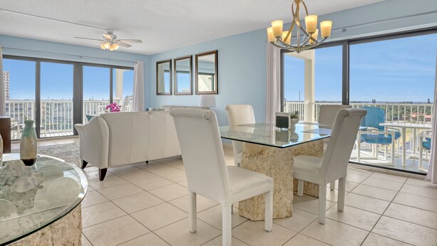 A modern living room with a glass dining table, white chairs, a sofa, and a view of the cityscape through large windows. - Stylish w/ Ocean View and 2 Balconies, Heated Pool, Centrally Located (Cocoa Beach)
