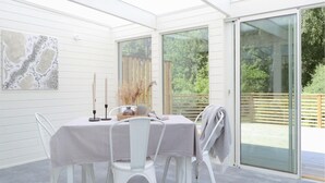 Un intérieur blanc, avec une table dressée pour deux personnes, un tableau accroché au mur et une porte-fenêtre coulissante donnant sur une terrasse extérieure.