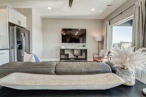 A modern living room with a grey sofa, a flat-screen TV, a kitchen area with a refrigerator, and a large window with curtains. - Epic 6BR Condo, Sleeps up to 24, Perfect for Your Group, Close to Everything! (Branson)