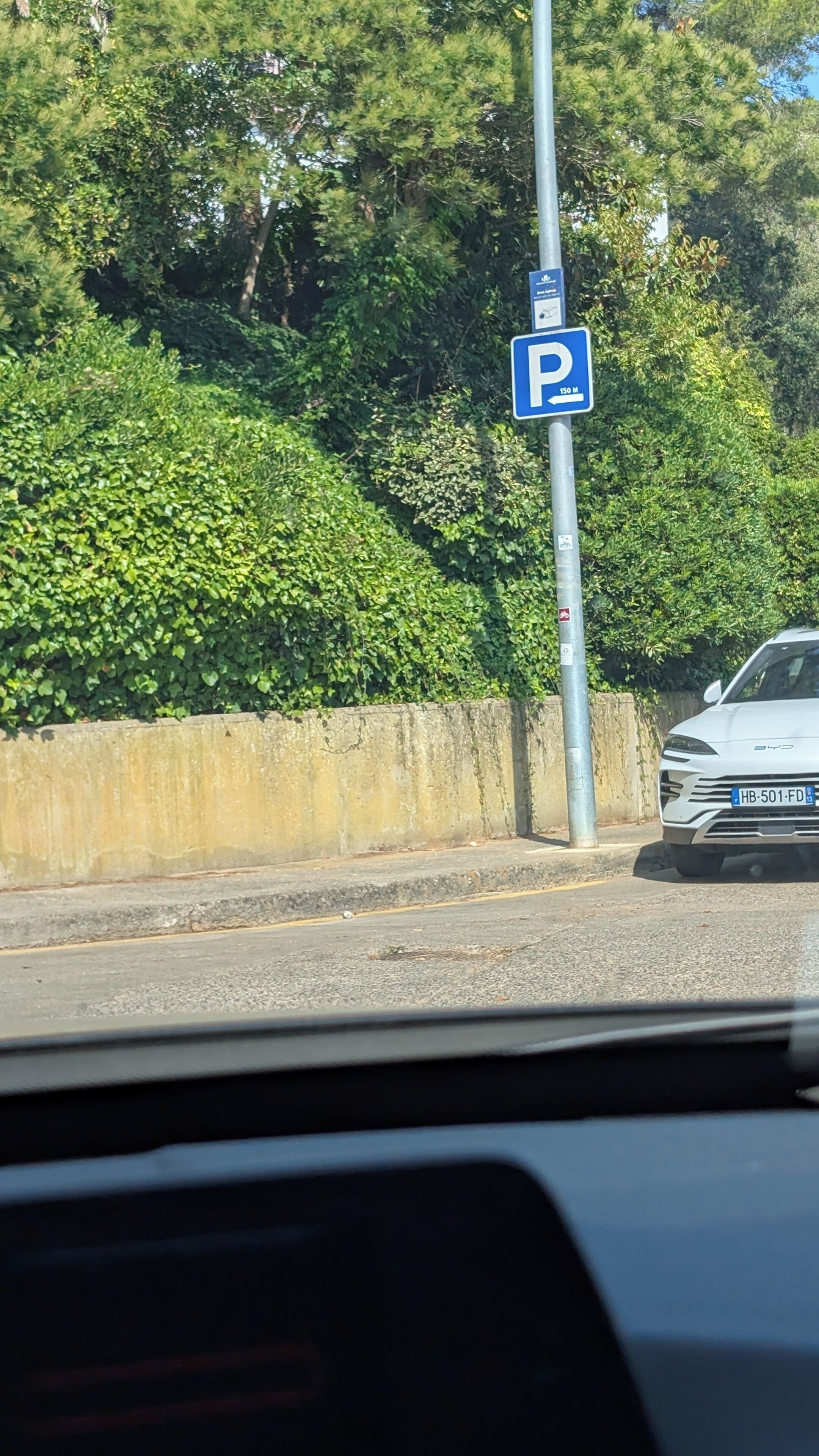 Una señal de estacionamiento con fondo azul y letras blancas, una barrera de concreto y un vehículo en la carretera.