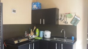 Een kleine keuken met een spoelbak, fornuis en kastjes. Bestek hangt aan de muur en er staat een doos bovenop de kastjes.