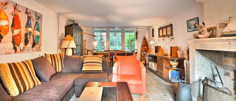 Een woonkamer met een bank, fauteuil en salontafel, een open haard, en een eethoek met tafel en stoelen.