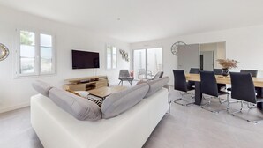 En moderne stue med en hvid sofa, et træsofabord og et tv ophængt på væggen. Der er et spiseområde med et træbord og stole, samt et stort spejl på væggen.