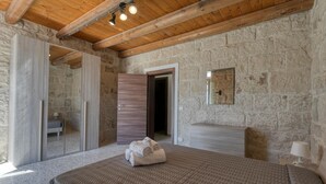 Ein Schlafzimmer mit Holzbalkendecke, Steinwänden, einem Bett mit gemustertem Überwurf, einem Nachttisch mit Lampe und einem Spiegel an der Wand.