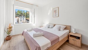 Un dormitorio con cama de madera, ropa de cama blanca, una mesita de noche, una lámpara de noche, una planta en maceta y una ventana con vistas a edificios.