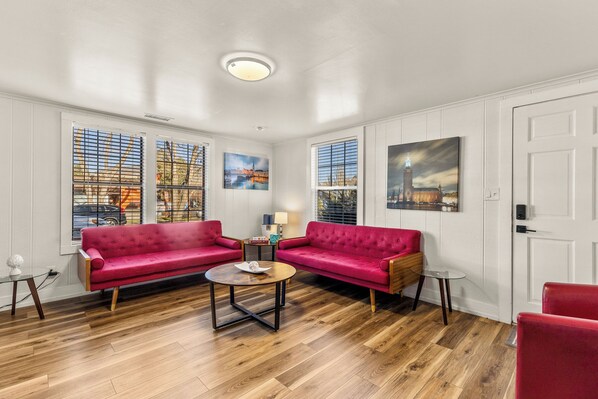 A spacious living room with two red sofas, a round coffee table, and a small side table. Large windows with blinds allow natural light and offer views of a cityscape.