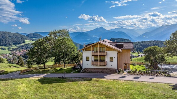 A two-story chalet with a balcony, surrounded by lush greenery and mountains.
