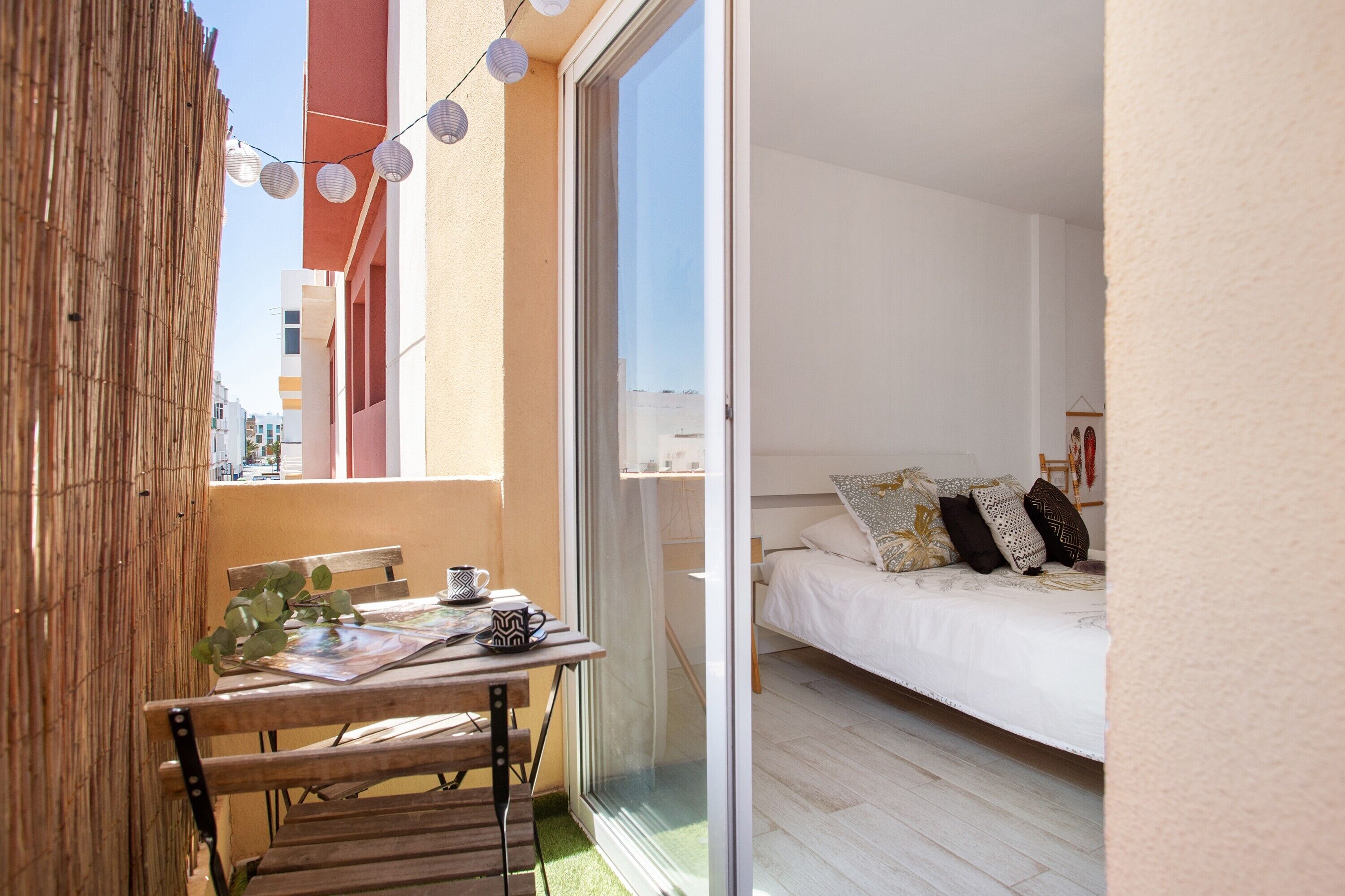 Un balcone con tavolo, sedie e luci a tendina; un letto con cuscini; e una porta scorrevole in vetro che dà su un ambiente interno.