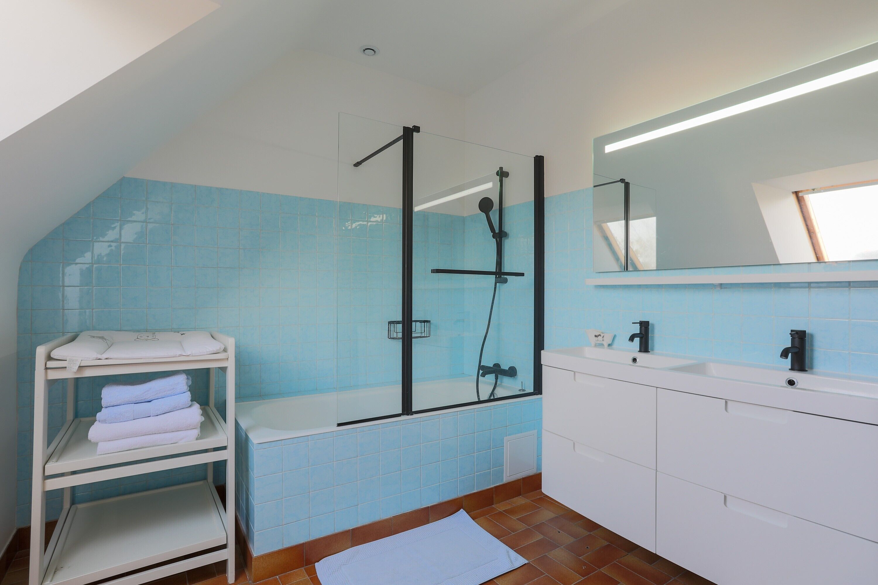 Une salle de bain avec une douche à parois de verre, des murs recouverts de carreaux blancs, un lavabo blanc et un porte-serviettes.
