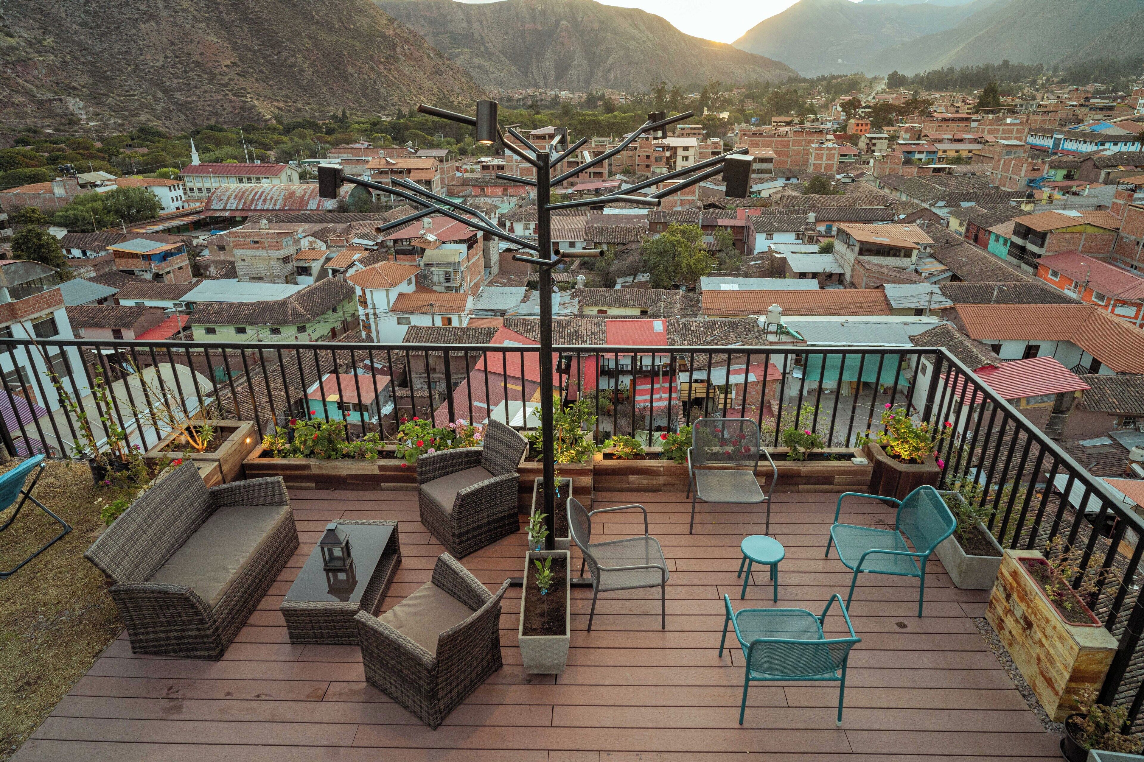 A deck with wicker furniture, a table, and chairs, overlooking a town with red-roofed buildings and mountains in the background.
