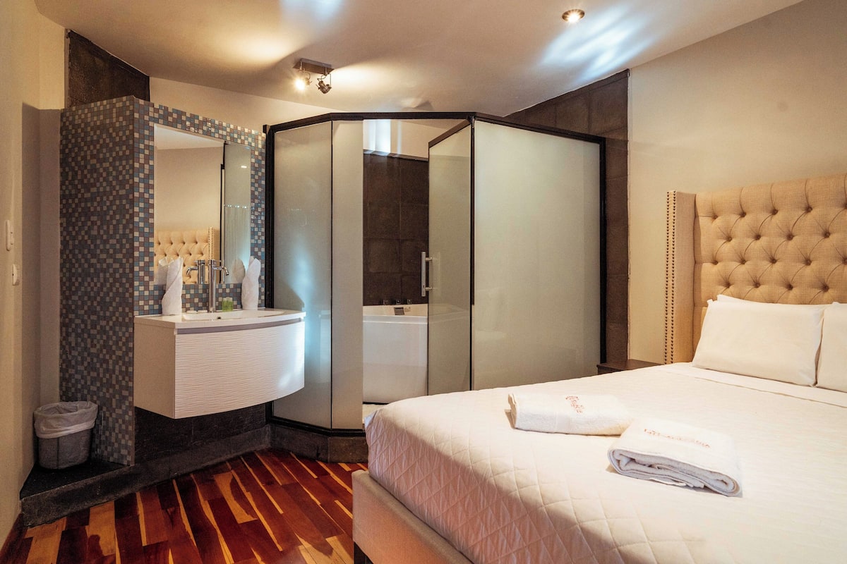 A hotel room with a large bed, a separate bathroom with a glass shower enclosure, and a vanity with a tiled backsplash.