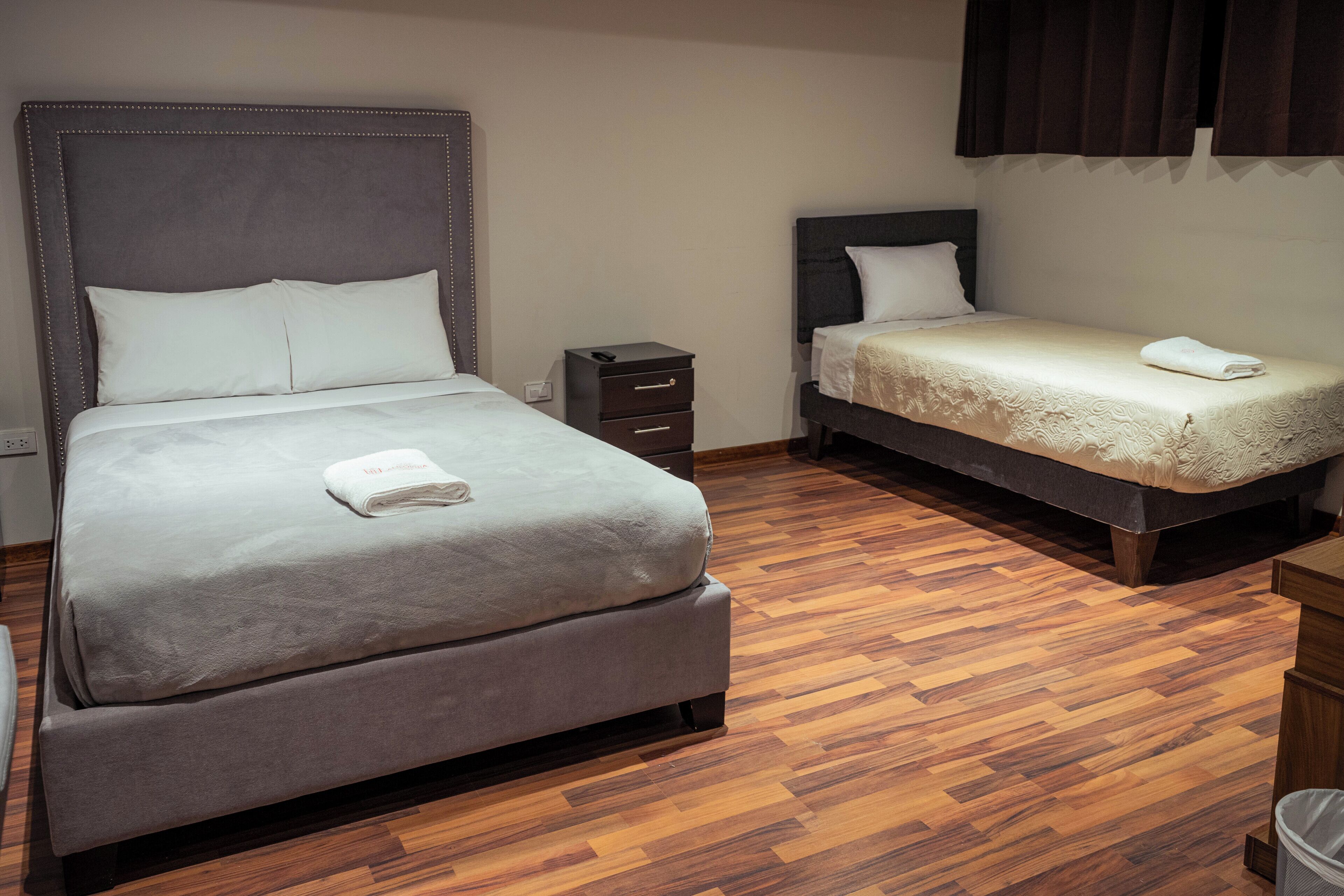 Una habitación de hotel con una cama grande, una cama más pequeña, una mesita de noche y piso de madera.