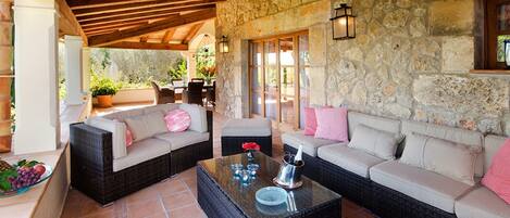 A covered outdoor seating area with wicker sofas, a glass coffee table, and stone walls.