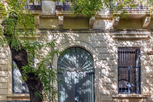 Fachada de edificio con un gran ventanal arqueado, una ventana con rejas y un árbol de follaje verde.