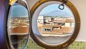 Vista a través de un ventanal redondo con marco de metal, que refleja un paisaje urbano con edificios y una montaña al fondo.