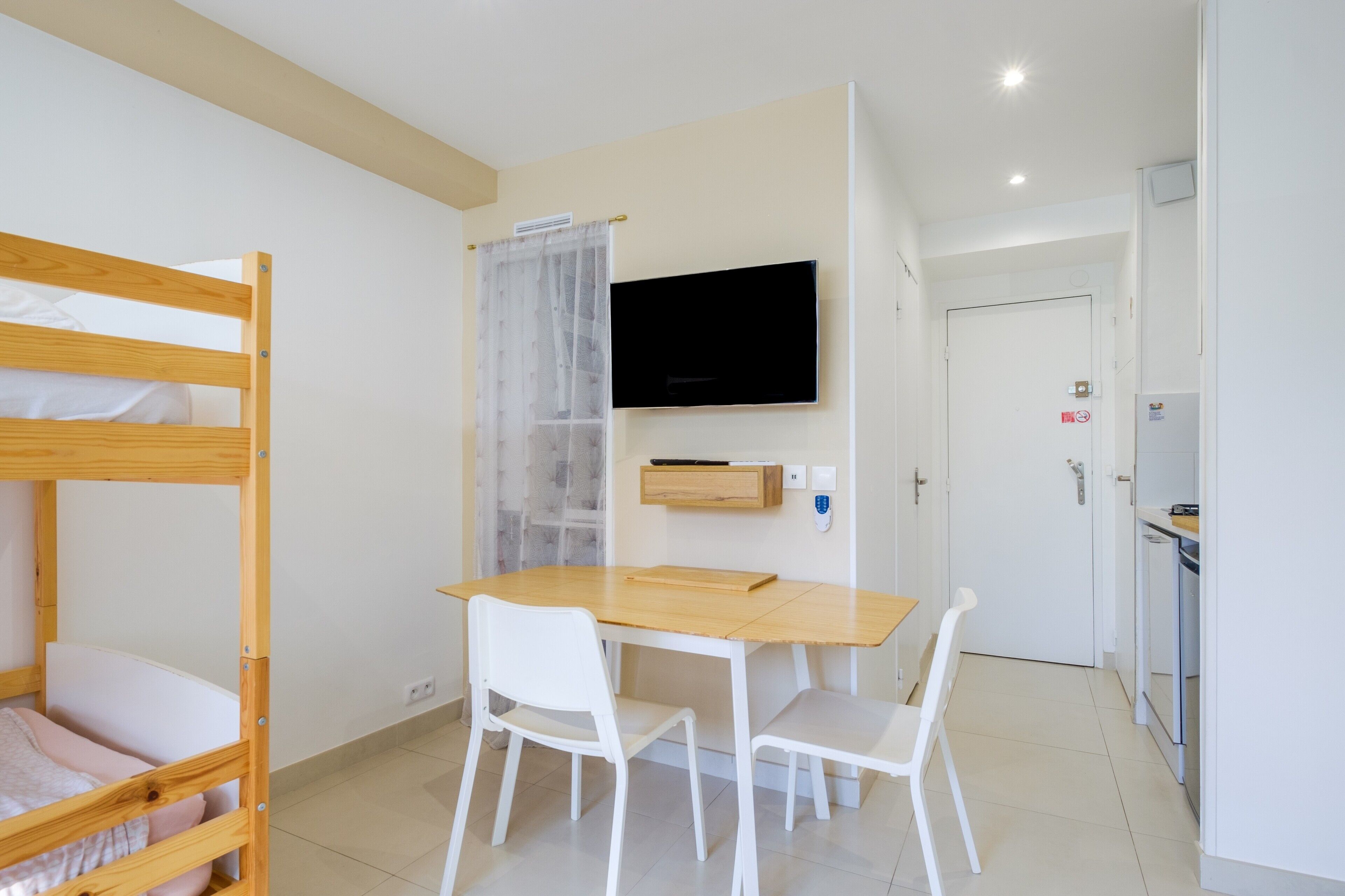 Una habitación con una cama litera, una mesa de madera con sillas, un televisor instalado en la pared y una puerta con manija.