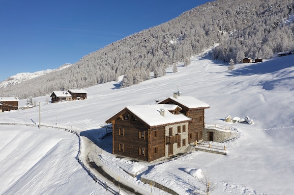 Eine Holzhütte mit schneebedecktem Dach, eingebettet in eine verschneite Landschaft und einen bewaldeten Berg.