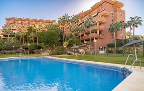 A swimming pool with a clear blue water surface, surrounded by a well-maintained lawn and palm trees. In the background, there is a multi-story building with balconies and a reddish-brown facade. - Lovely apartment in Benalmádena (Benalmádena)