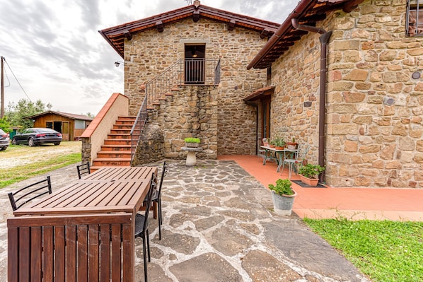 A stone building with a wooden staircase leading to a balcony, outdoor seating, and potted plants.