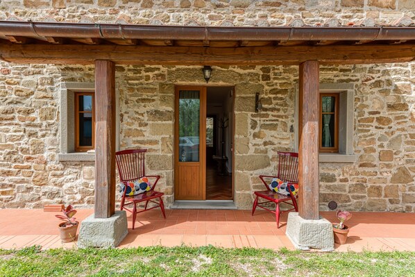 Un bâtiment en pierre avec un porche en bois, deux chaises rouges et une porte donnant sur l’intérieur.