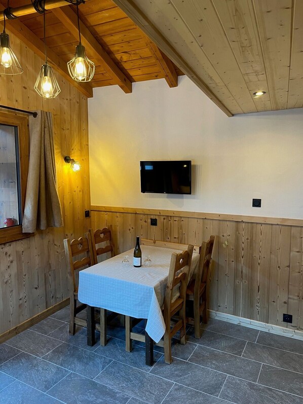 Un coin repas avec une table dressée pour deux personnes, des chaises en bois, une bouteille de vin et un téléviseur à écran plat fixé au mur.