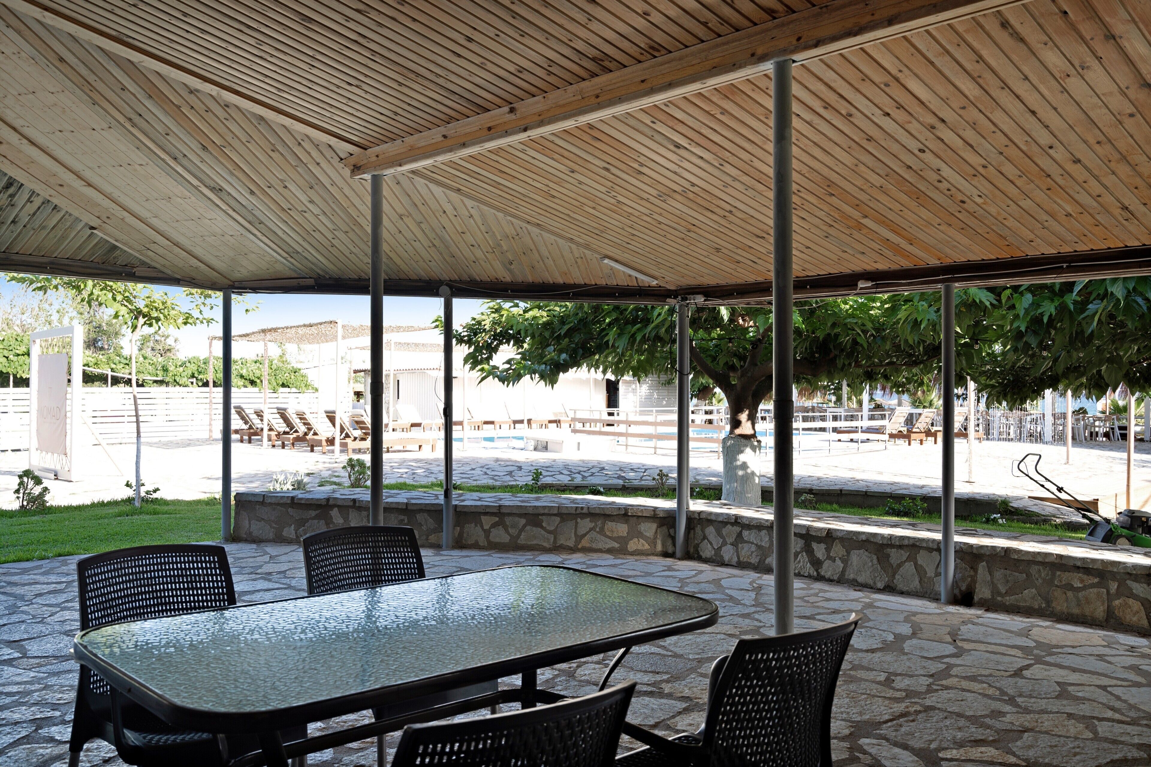 An outdoor patio with a table and chairs, a wooden ceiling, and a view of a pool and greenery.