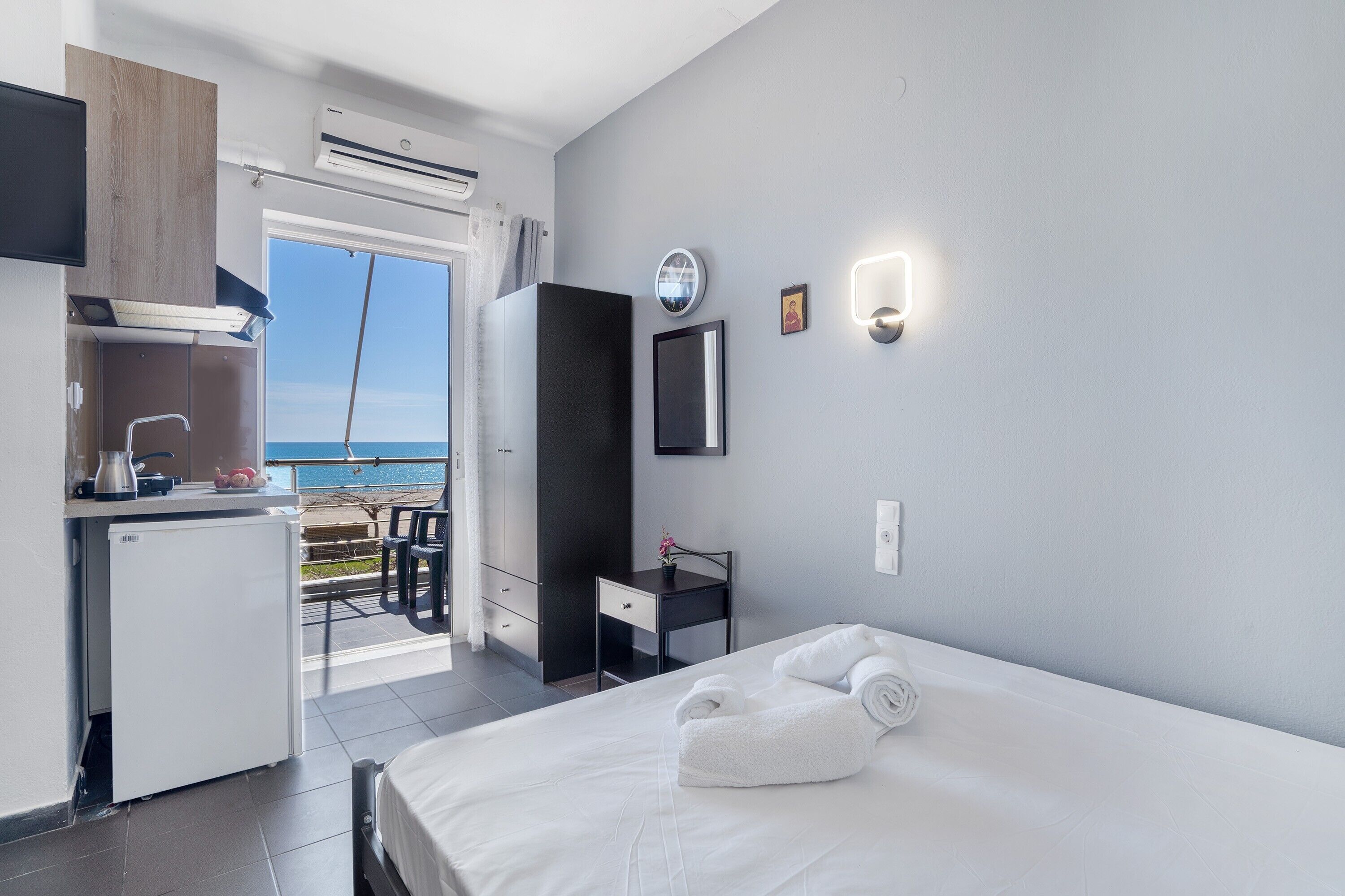 Habitación de hotel con cama, escritorio, kitchenette pequeña, balcón con vista al mar y un reloj de pared.