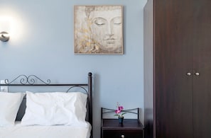 A bedroom with a bed, white pillows, a nightstand with a flower, a wall-mounted artwork, and a wardrobe.