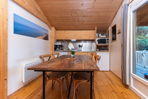 Une table à manger en bois avec des chaises, un radiateur blanc, des meubles en bois, un coin cuisine équipé d’appareils électroménagers et une fenêtre avec vue.