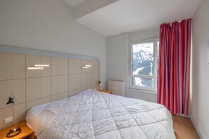 A bedroom with a bed, a nightstand, a lamp, a window with curtains, and a view of mountains.