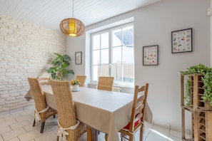 Un coin repas avec une table blanche, des chaises en osier, une lampe suspendue et une fenêtre avec des stores.