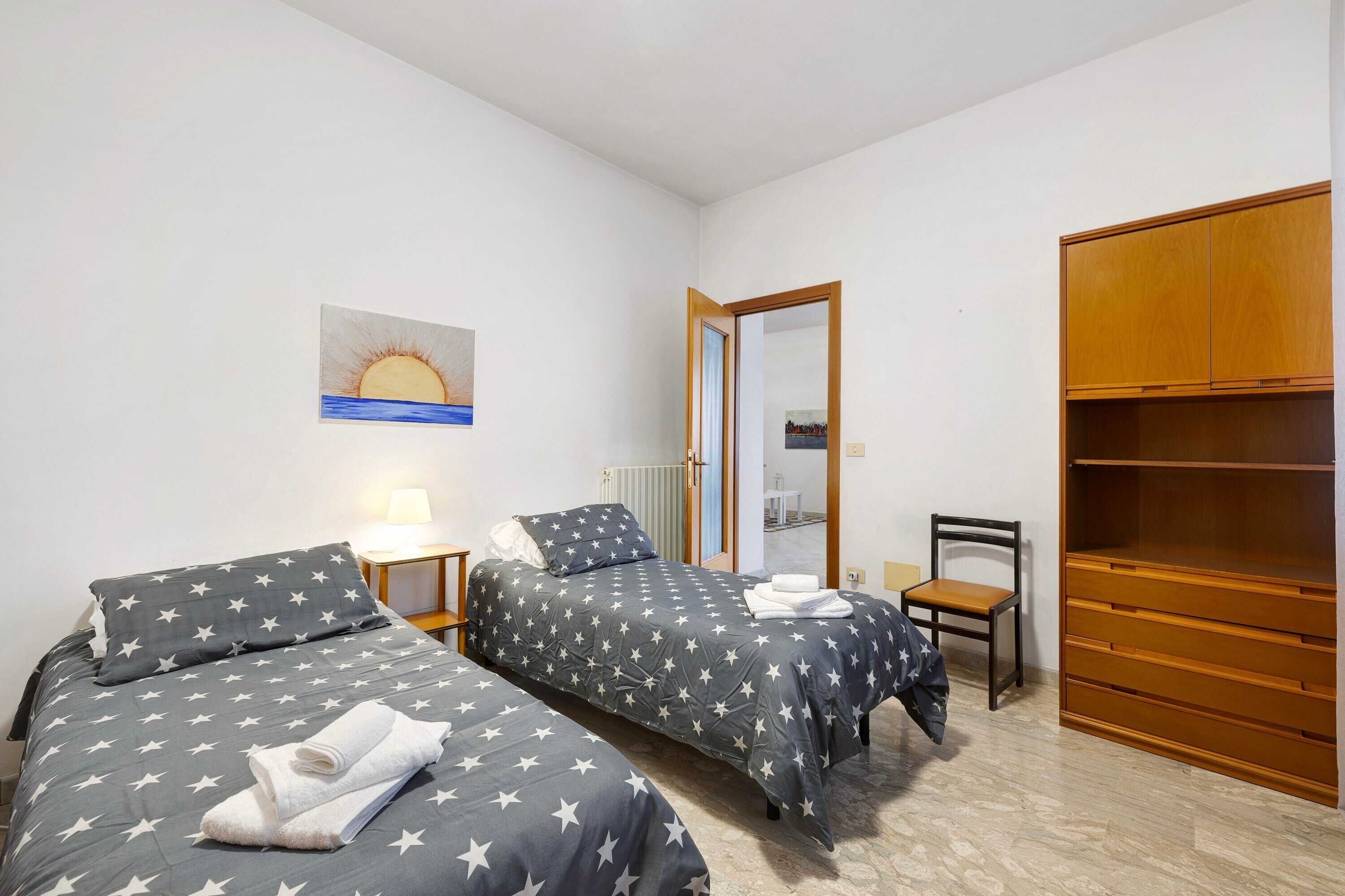 A bedroom with two beds, a wooden wardrobe, a chair, a desk, and a painting on the wall.