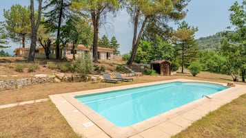 Une piscine entourée d’arbres et d’un mur de pierre, avec des chaises longues à proximité.