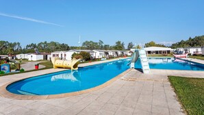 A swimming pool with a slide, surrounded by a paved area and mobile homes in the background.