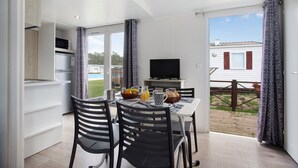 A dining area with a table set for two, a television, and a kitchen area with a microwave and refrigerator. - Chalet 'Bungalow T2 I V- 15 Min From Nazaré' with Shared Pool, Private Terrace and Air Conditioning (Pataias)