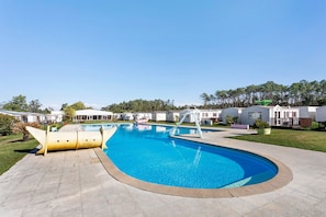 Uma piscina com escorrega, rodeada por um espaço pavimentado e casas pequenas.