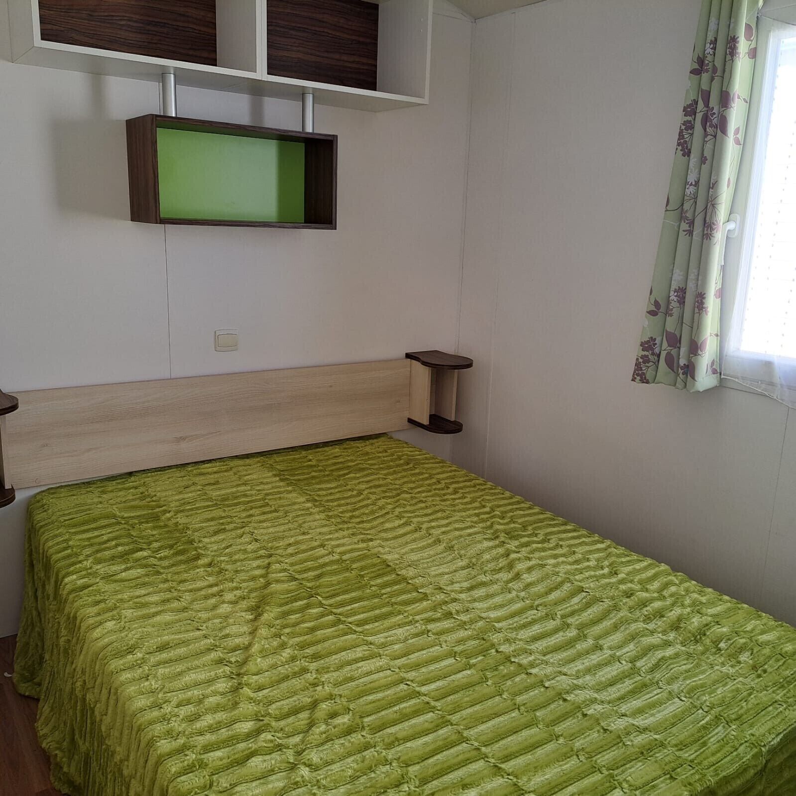 Un lit simple avec une couette verte, une tête de lit en bois, une table de chevet et un téléviseur fixé au mur.
