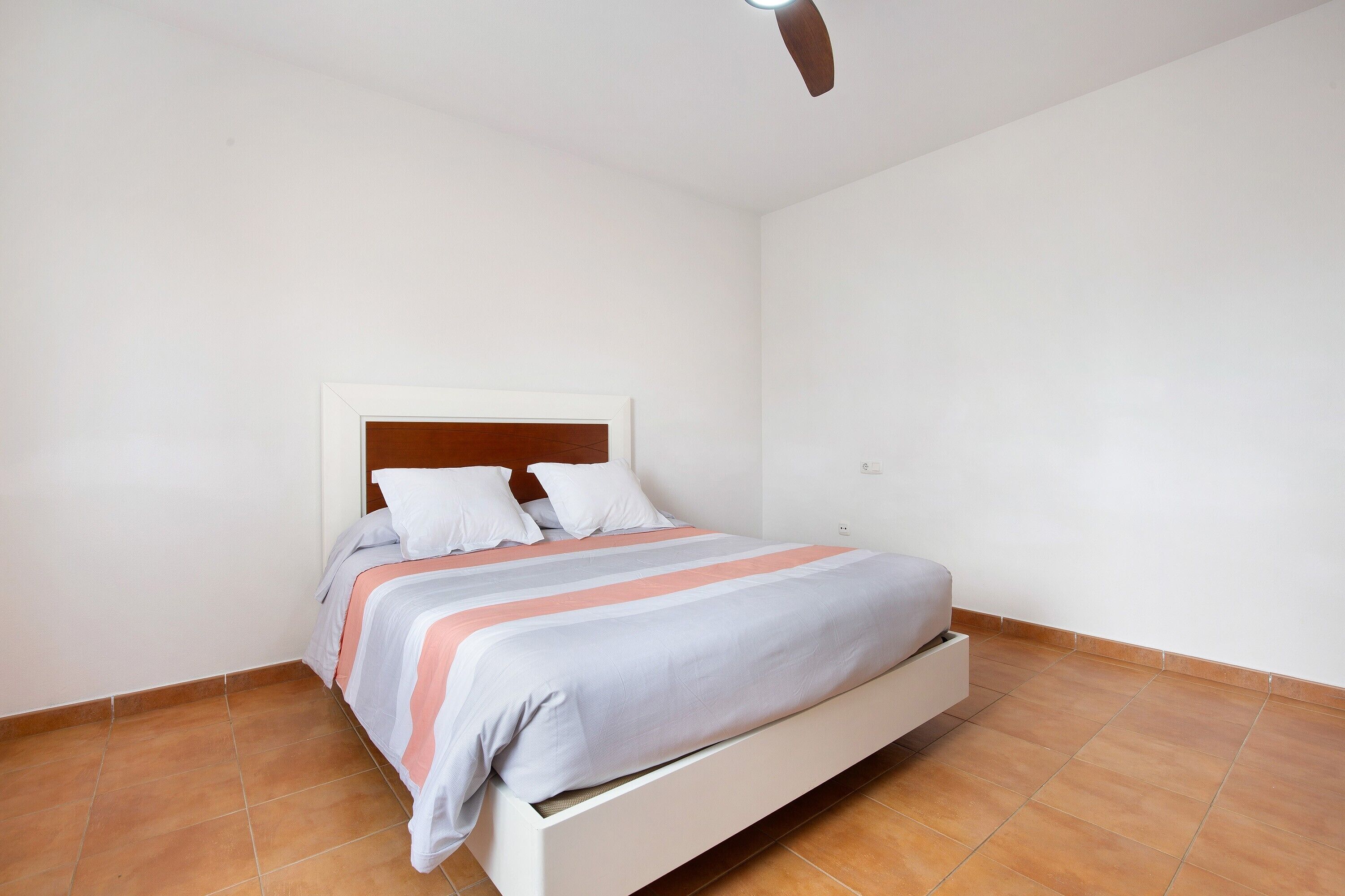 Un lit simple avec une tête de lit en bois, des murs blancs et un sol carrelé.