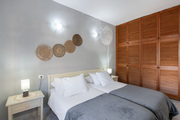 Een tweepersoonsbed met wit beddengoed, een nachtkastje met een lamp, een wandklok en een houten garderobe.