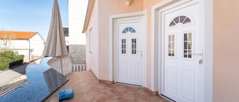 A clean, modern exterior of a building with two white doors, a small patio area with a blue countertop and a beige umbrella, and a tiled floor.