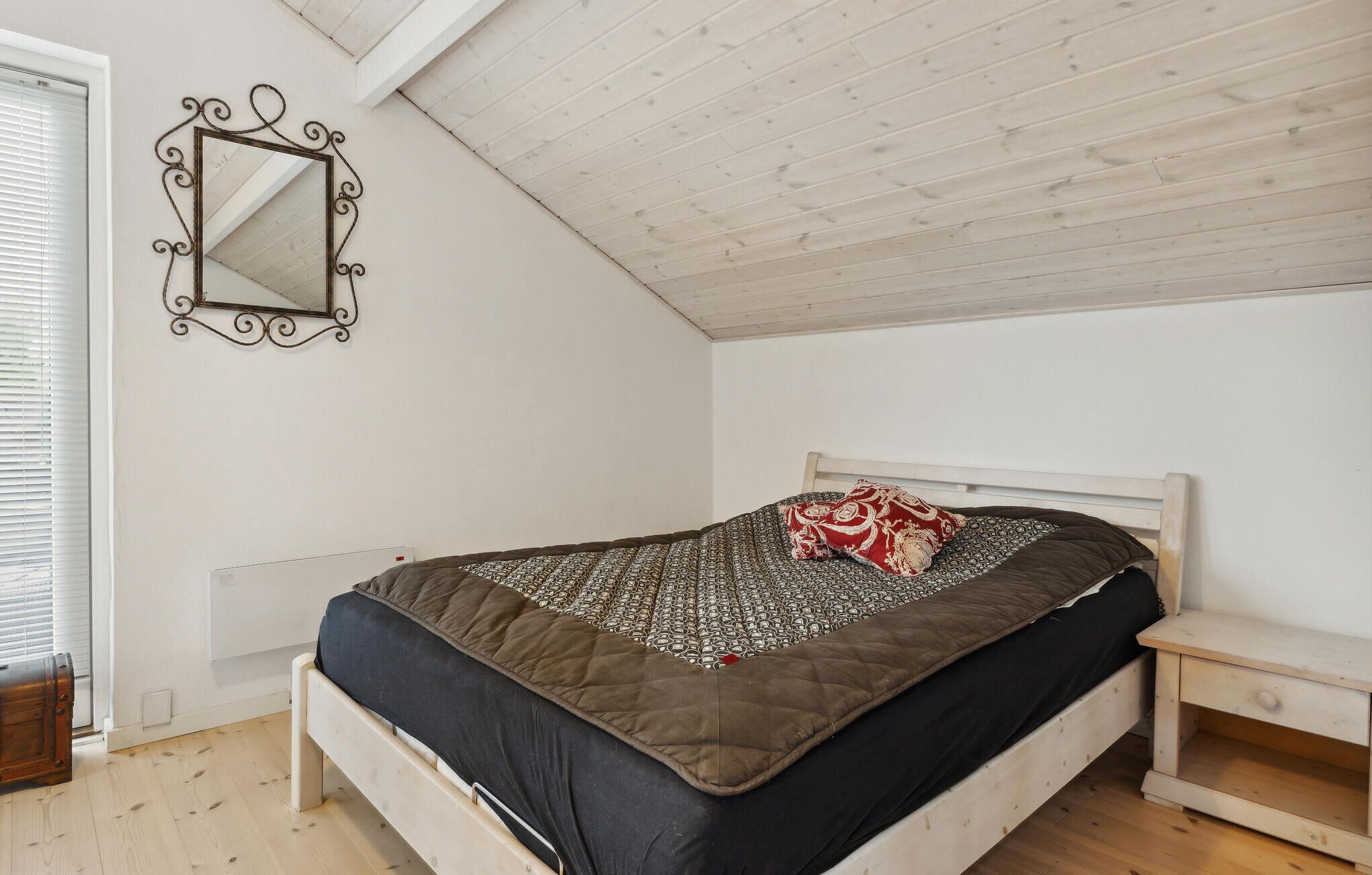 A bedroom with a bed, a bedside table, a nightstand, a mirror, and a window with blinds.