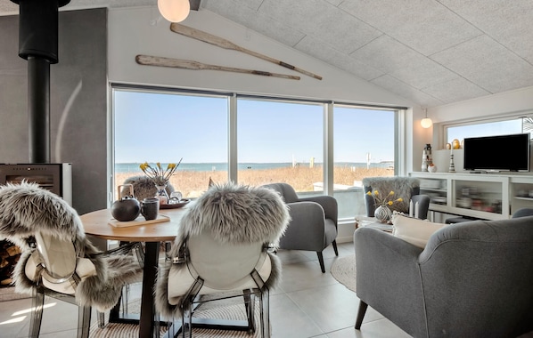 A modern living room with a large window overlooking a beach, a fireplace, and a television on a white console.